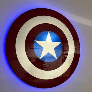 Puede incluir: Un escudo de Capitán América rojo, blanco y azul con una luz LED azul alrededor del borde. El escudo está montado en una pared blanca.