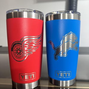 May include: Two insulated tumblers. One is red with a silver Detroit Red Wings logo. The other is blue with a silver Detroit Lions logo. Both have a silver rim and the Yeti brand name.