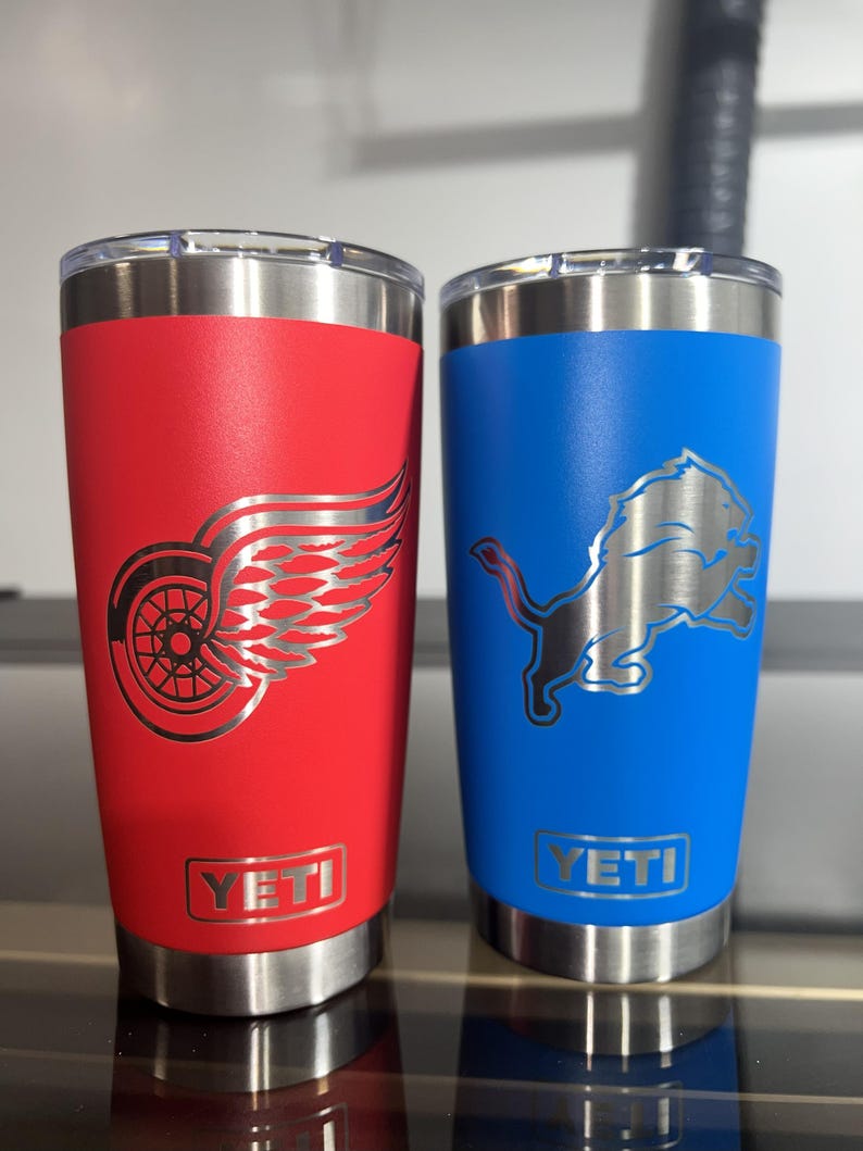 May include: Two insulated tumblers. One is red with a silver Detroit Red Wings logo. The other is blue with a silver Detroit Lions logo. Both have a silver rim and the Yeti brand name.