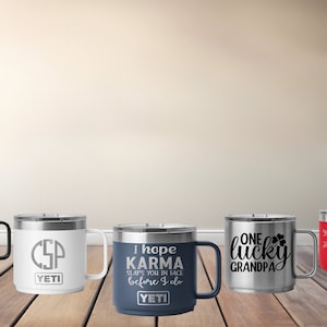 Op de afbeelding: Vijf Yeti-merk reistassen van roestvrij staal met verschillende ontwerpen en kleuren. De bekers staan op een houten oppervlak. De ontwerpen omvatten "Land of the Free, Home of the Brave", "CSP", "Ik hoop dat Karma je in je gezicht slaat voordat ik het doe", "Een gelukkige opa" en een bloemenontwerp.