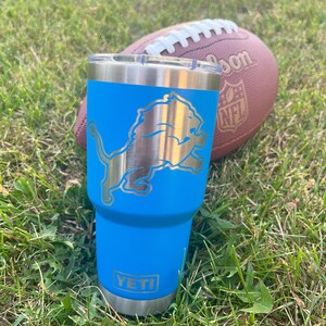 May include: A blue Yeti tumbler with a silver Detroit Lions logo and the word "YETI" on the bottom. A brown football with white laces rests in the background on a bed of green grass.