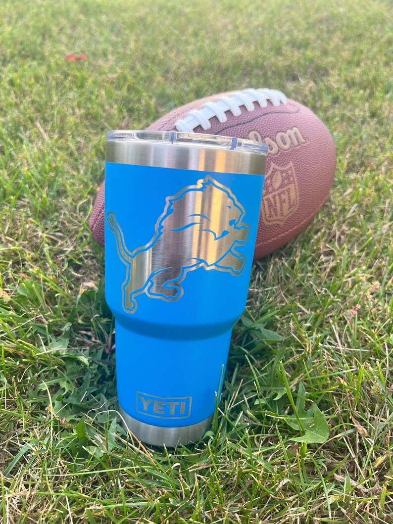 May include: A blue Yeti tumbler with a silver Detroit Lions logo and the word "YETI" on the bottom. A brown football with white laces rests in the background on a bed of green grass.