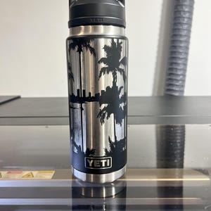 May include: A stainless steel YETI water bottle with a black handle and lid. The bottle features a black silhouette design of palm trees and a city skyline. The YETI logo is visible on the bottle.