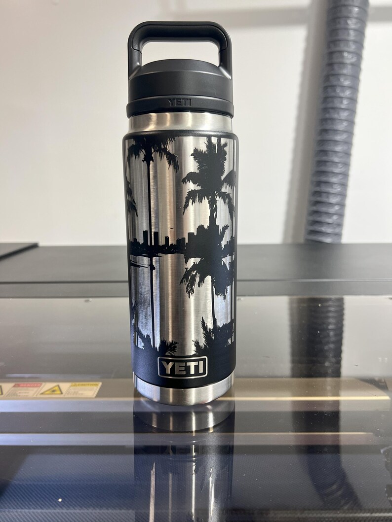 May include: A stainless steel YETI water bottle with a black handle and lid. The bottle features a black silhouette design of palm trees and a city skyline. The YETI logo is visible on the bottle.
