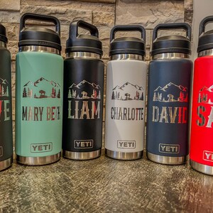 May include: Six insulated water bottles in black, seafoam green, black, white, navy blue, and red. Each bottle has a mountain and tree design with a name. The brand name "YETI" is visible.