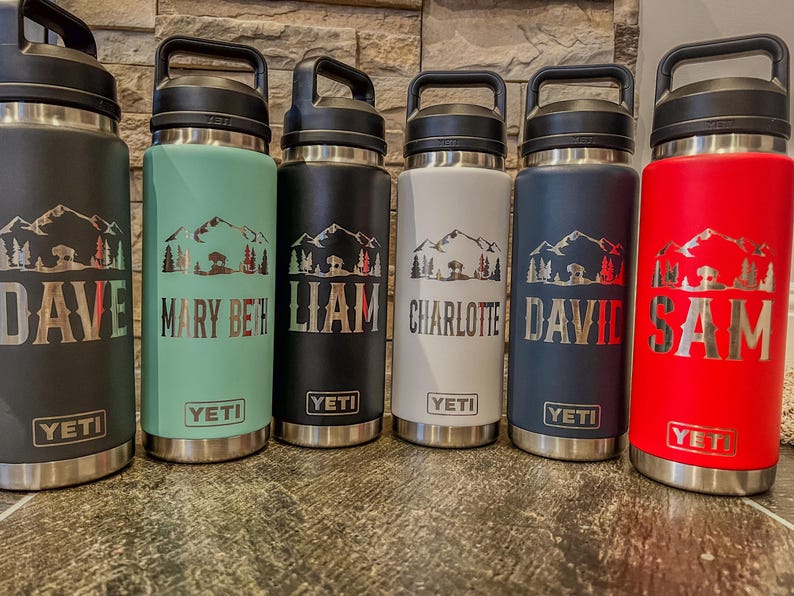 May include: Six insulated water bottles in black, seafoam green, black, white, navy blue, and red. Each bottle has a mountain and tree design with a name. The brand name "YETI" is visible.