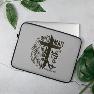 Puede incluir: Funda para portátil gris con un diseño de león negro y cruz. El texto "Man of Faith" y "1 Corintios 16:13" está impreso en la funda.