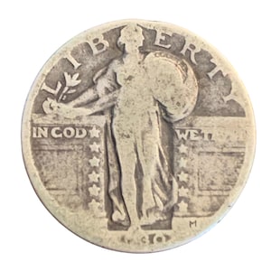 May include: A silver Standing Liberty quarter coin. The coin features the word "LIBERTY" at the top, and the words "IN GOD WE TRUST" are visible. The coin has a depiction of a woman holding a branch and shield.