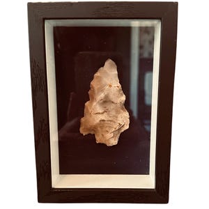 Puede incluir: Una punta de flecha enmarcada, probablemente de piedra, en tonos marrón claro y beige. La punta de flecha se exhibe en un marco de madera marrón oscuro con un borde blanco, sobre un fondo oscuro.