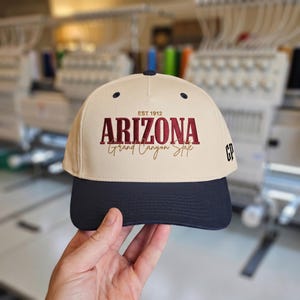 Könnte beinhalten: Eine beige Baseballkappe mit dunkelblauem Schirm und oberem Knopf. Die Vorderseite zeigt in Dunkelrot und Gold gestickten Text mit der Aufschrift EST 1912 ARIZONA Grand Canyon State. An der Seite befinden sich die schwarz gestickten Buchstaben GP.