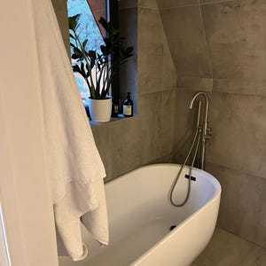 May include: A modern bathroom with a white oval bathtub and a silver faucet. A white towel hangs near the tub. A potted plant sits in a window, and the walls are tiled in gray.