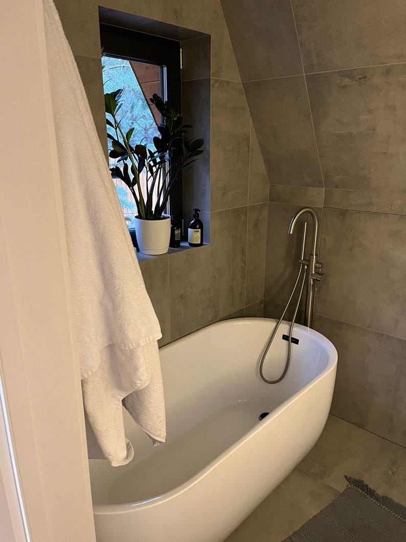 May include: A modern bathroom with a white oval bathtub and a silver faucet. A white towel hangs near the tub. A potted plant sits in a window, and the walls are tiled in gray.