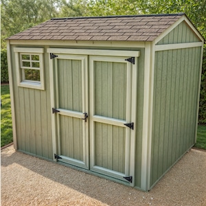 May include: A small, green wooden shed with a brown shingled roof. The shed has a small window and double doors with black hinges and a latch. The trim is a light cream colour. The shed is on a gravel surface.