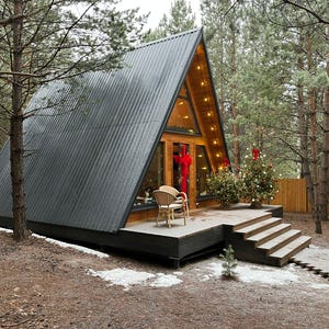 May include: A-frame cabin with a dark gray corrugated metal roof and large glass windows. The wooden deck has a small Christmas tree and a wicker chair. The cabin is surrounded by trees and a wooden fence.