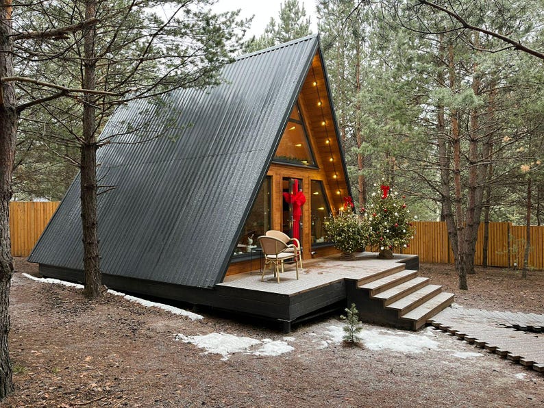 May include: A-frame cabin with a dark gray corrugated metal roof and large glass windows. The wooden deck has a small Christmas tree and a wicker chair. The cabin is surrounded by trees and a wooden fence.