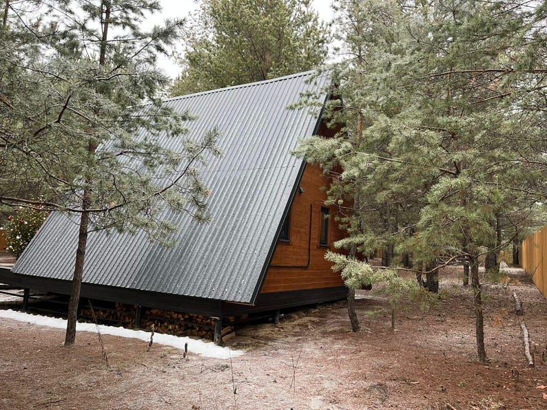 May include: A-frame cabin with a corrugated metal roof nestled among pine trees. The wooden structure has a dark brown exterior and sits on a raised foundation. Firewood is stacked beneath the cabin, and snow covers the ground.