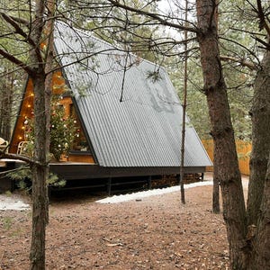 May include: An A-frame cabin with a gray corrugated metal roof is nestled among tall pine trees. A Christmas tree is visible inside the cabin. The cabin has a wooden deck and is surrounded by a forest floor of pine needles.