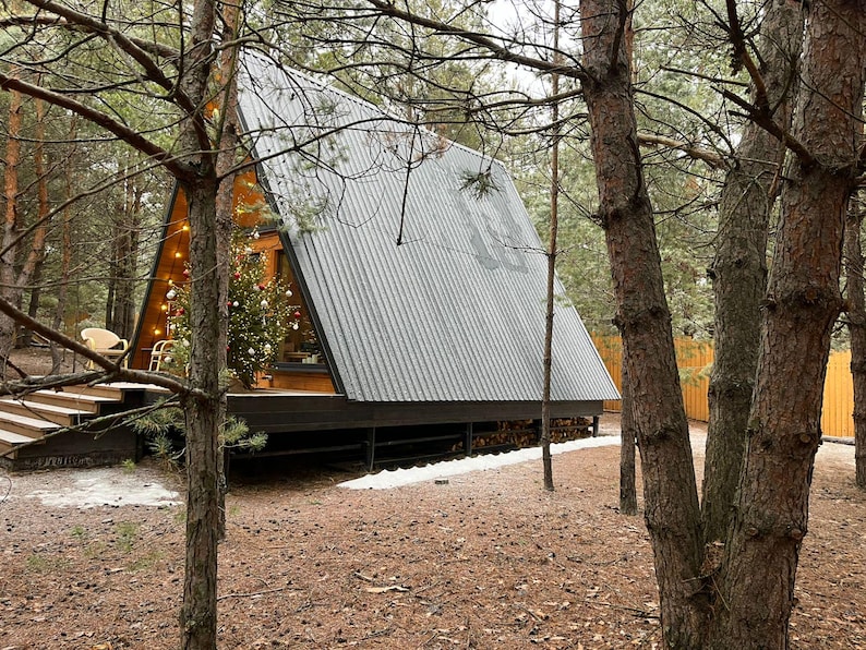 May include: An A-frame cabin with a gray corrugated metal roof is nestled among tall pine trees. A Christmas tree is visible inside the cabin. The cabin has a wooden deck and is surrounded by a forest floor of pine needles.