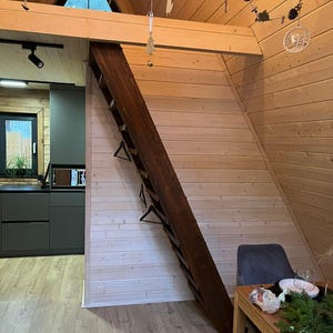 May include: Interior shot of a cabin with wooden walls and ceiling. A dark brown wooden ladder leans against a light-colored wall. A small kitchen with dark gray cabinets is visible. A dining table with a green garland is set up.