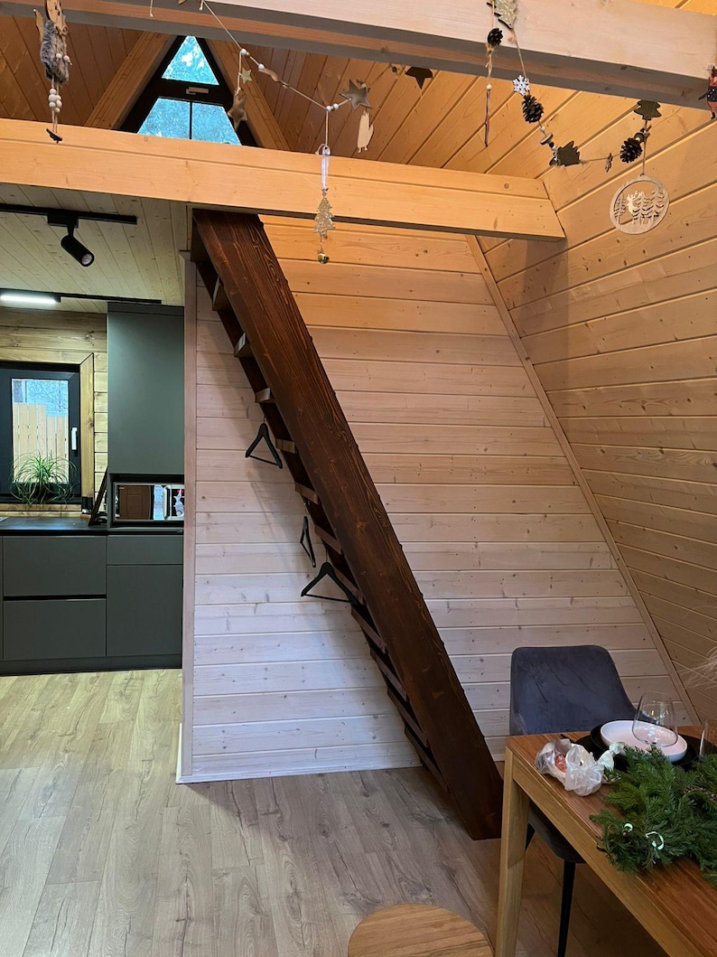 May include: Interior shot of a cabin with wooden walls and ceiling. A dark brown wooden ladder leans against a light-colored wall. A small kitchen with dark gray cabinets is visible. A dining table with a green garland is set up.