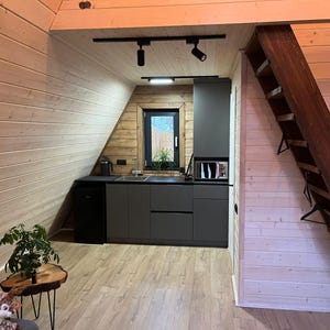 May include: Interior shot of a small kitchen with gray cabinets, a microwave, and a small refrigerator. The kitchen is built into an angled wall with wood paneling. A wooden staircase leads to an upper level. A small plant sits on a wooden table.