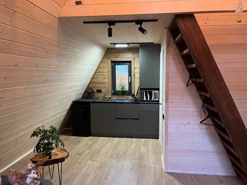May include: Interior shot of a small kitchen with gray cabinets, a microwave, and a small refrigerator. The kitchen is built into an angled wall with wood paneling. A wooden staircase leads to an upper level. A small plant sits on a wooden table.
