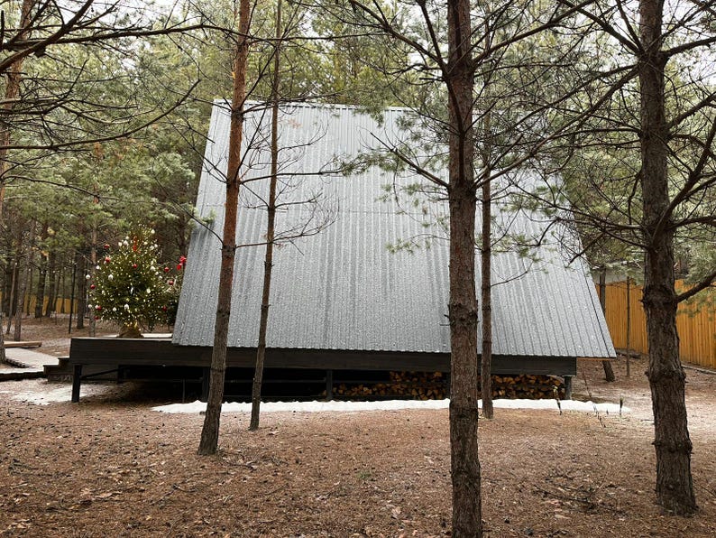 May include: A-frame cabin with a corrugated metal roof, nestled among pine trees. A Christmas tree is visible in the background. The cabin sits on a dark wooden deck, with firewood stored underneath. The ground is covered in fallen leaves and a dusting of snow.
