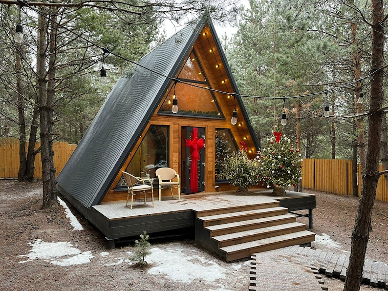May include: A-frame cabin with a dark gray corrugated metal roof and wooden siding. The cabin has large windows and a small porch with steps. A Christmas tree is decorated on the porch, and string lights are strung around the cabin.