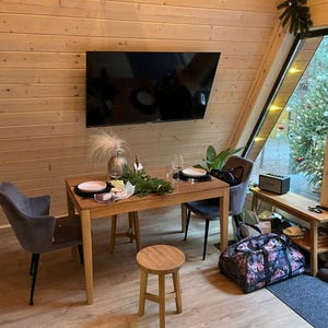 May include: A cozy dining area with a wooden table set for a meal. Two gray chairs are positioned around the table, and a small wooden stool sits nearby. A large TV is mounted on the wall, and a window offers a view of a decorated Christmas tree.