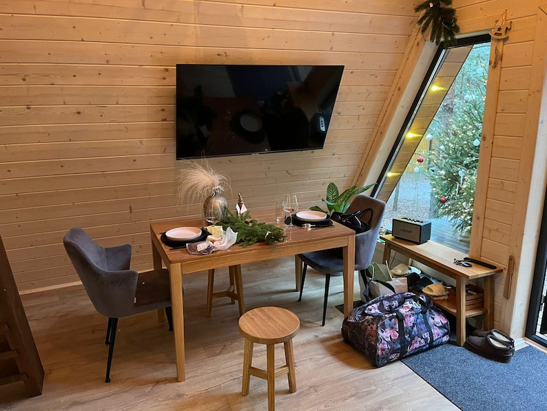 May include: A cozy dining area with a wooden table set for a meal. Two gray chairs are positioned around the table, and a small wooden stool sits nearby. A large TV is mounted on the wall, and a window offers a view of a decorated Christmas tree.