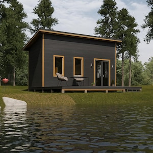 Puede incluir: Una cabaña gris oscuro con una terraza de madera y molduras doradas se encuentra a la orilla de un lago. Dos sillas y una mesa pequeña están en la terraza. Altos árboles verdes rodean la cabaña y una parrilla roja está en el césped.
