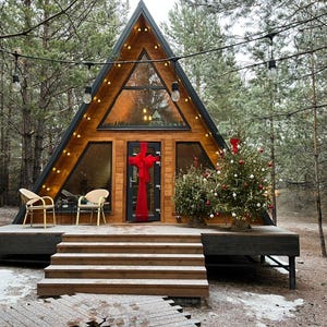 May include: A-frame cabin with a wooden exterior, large windows, and a black roof. The cabin is decorated for the holidays with a Christmas tree and a red bow on the door. Two chairs sit on the deck.