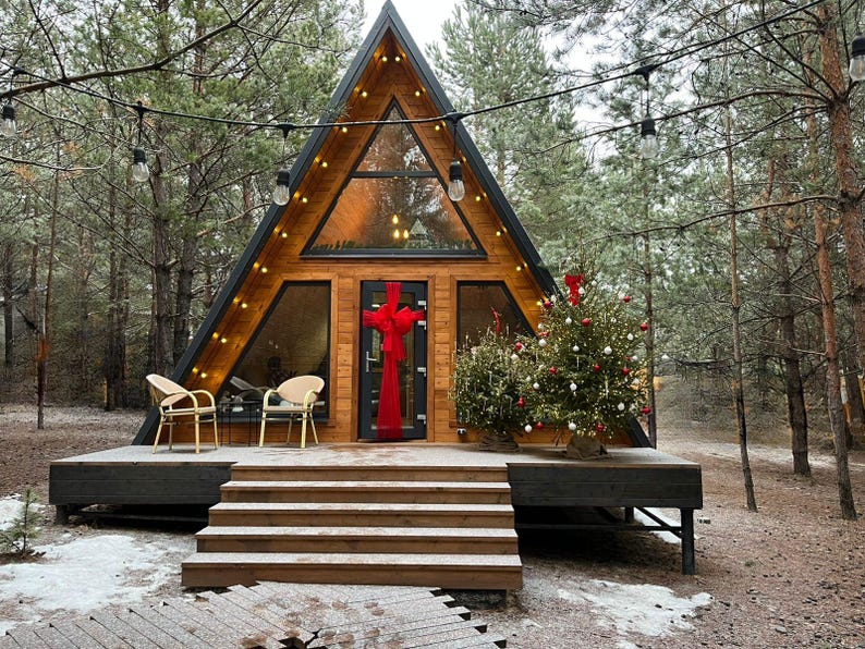 May include: A-frame cabin with a wooden exterior, large windows, and a black roof. The cabin is decorated for the holidays with a Christmas tree and a red bow on the door. Two chairs sit on the deck.