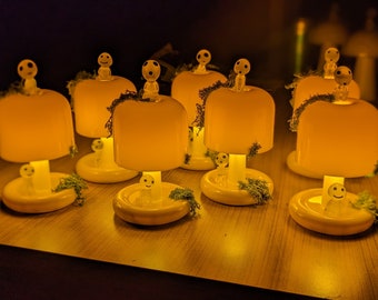 May include: A group of seven small, yellow ceramic lamps with white figures on top. Each lamp has a small green base and a white figure with a smiling face. The lamps are lit and casting a warm glow.