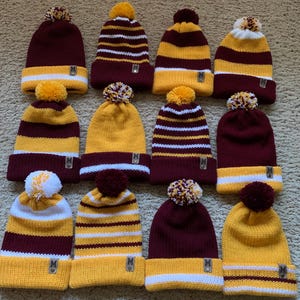 Puede incluir: Una colección de gorros de punto en varias combinaciones de colores. Los gorros tienen un pompón en la parte superior y una pequeña etiqueta con la letra "M". Los esquemas de color incluyen granate, dorado y rayas blancas.