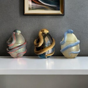 May include: Three decorative egg-shaped containers with snake detailing. One features a silver and red snake, another a gold snake, and the third a blue snake. The containers are on a white shelf against a grey wall.