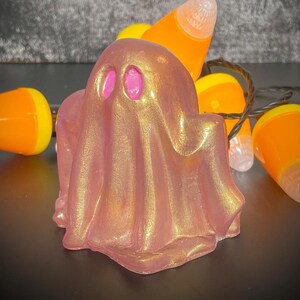 May include: A pink and gold ghost figurine with pink eyes. The ghost is surrounded by candy corn lights, creating a festive Halloween theme. The figurine has a shimmering, metallic finish.