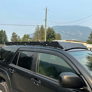 May include: A black SUV with a black metal roof rack. The roof rack has a flat, rectangular design. The vehicle is parked outdoors on a sunny day, with trees and mountains visible in the background.