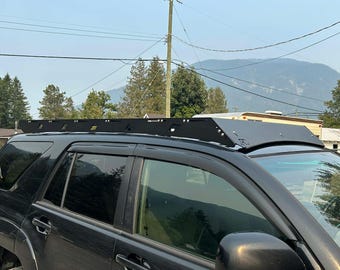 Könnte beinhalten: Ein schwarzer SUV mit einem schwarzen Metalldachgepäckträger. Der Dachträger hat ein flaches, rechteckiges Design. Das Fahrzeug steht an einem sonnigen Tag im Freien, mit Bäumen und Bergen im Hintergrund.
