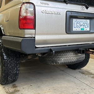 Puede incluir: Vista trasera de un Toyota 4Runner beige con neumáticos negros grandes. El vehículo tiene un parachoques negro y un neumático de repuesto montado debajo. Se ve una placa de matrícula.