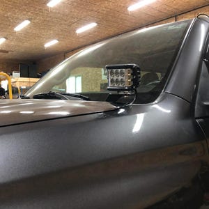 以下が含まれることがあります： グレーの車両のボンネットに取り付けられた黒色の LED ライトバー。 ライトバーには、複数の小さな正方形のライトがあります。 車両は、上部照明のあるガレージの中に駐車されています。