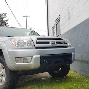 May include: A silver Toyota 4Runner SUV with a black front bumper and a V6 emblem on the front grille. The vehicle is parked in front of a white brick wall.