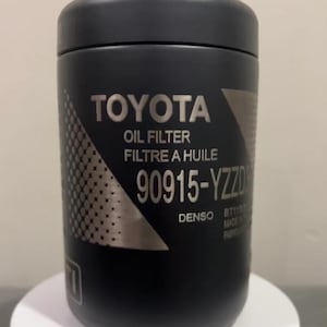 Könnte beinhalten: Ein schwarzer Ölfilter mit silberner Beschriftung "TOYOTA OIL FILTER FILTRE A HUILE 90915-YZZD DENSO BT1168K MADE IN JAPAN FABRIQUE AU JAPON".