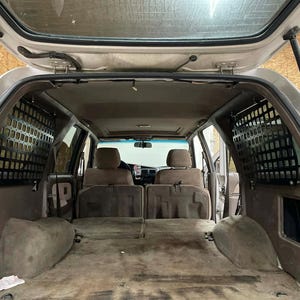 May include: Interior view of a light-coloured SUV with the rear hatch open. The cargo area features brown carpeting and side panels, with black metal mesh dividers. The seats are a matching brown colour. The vehicle's roof is visible.