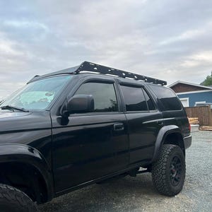 Könnte beinhalten: Schwarzer SUV mit Dachgepäckträger, geparkt auf einer Schotterauffahrt. Das Fahrzeug hat große Geländereifen und getönte Scheiben. Der Himmel ist bewölkt, und im Hintergrund sind Häuser zu sehen.