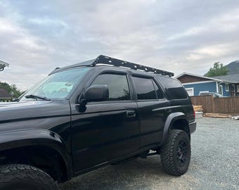 Könnte beinhalten: Schwarzer SUV mit Dachgepäckträger, geparkt auf einer Schotterauffahrt. Das Fahrzeug hat große Geländereifen und getönte Scheiben. Der Himmel ist bewölkt, und im Hintergrund sind Häuser zu sehen.