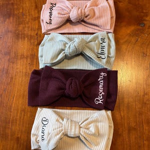 May include: Four ribbed headbands in pastel colors: pink, sage green, burgundy, and cream. Each headband features a large bow and a name in cursive script: Rosemary, Elanora, Rosemary, and Diana. The headbands are displayed on a wooden surface.