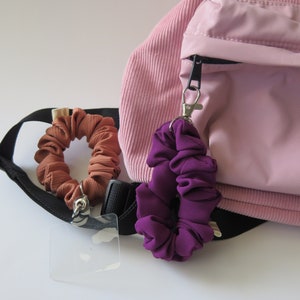 May include: A pink corduroy backpack with a black zipper and a purple and brown scrunchie attached to the zipper pull.