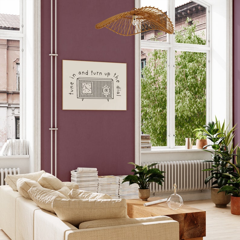 May include: A black and white illustration of a vintage radio with the text "tune in and turn up the dial" on a cream background. The illustration is framed and hanging on a burgundy wall.