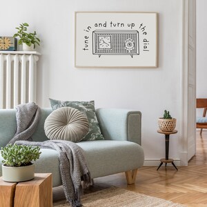 May include: A black and white illustration of a vintage radio with the text "tune in and turn up the dial". The illustration is framed and hanging on a white wall above a light blue sofa with a throw blanket and pillows.